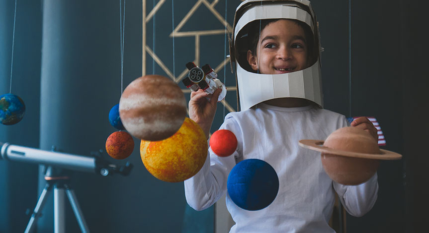 boy wearing cardboard astronaut helmet flying toy rocket through planets, cardboard spaceship rocket in the background